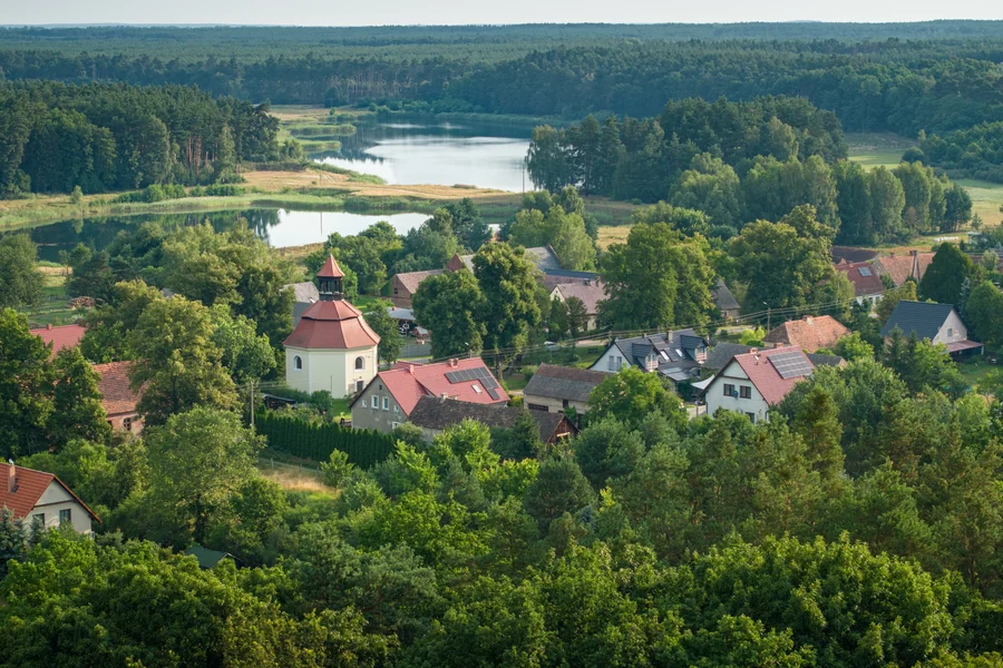 Wieś z niewielkim kościołem wśród drzew, w tle dwa jeziora i las, widok z lotu ptaka