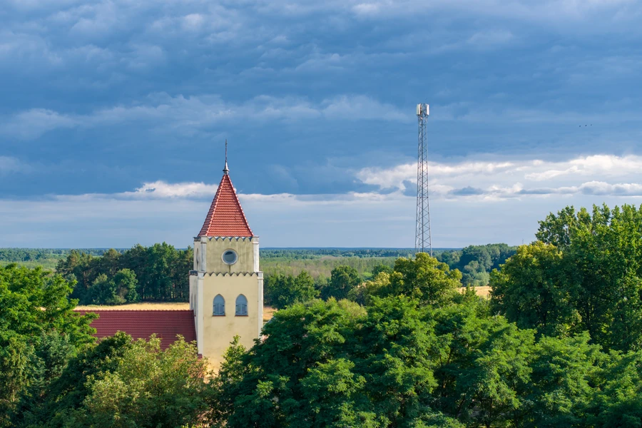 Wieża kościoła zwieńczona czerwonym hełmem wśród drzew, w tle maszt i pola, widok z lotu ptaka