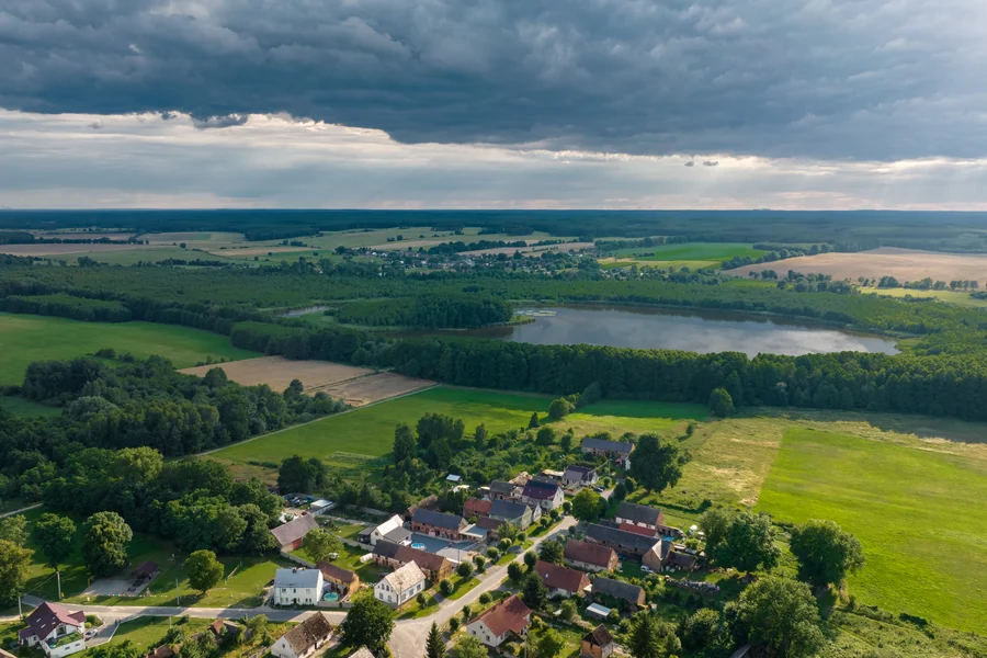 Zabudowa wsi wśród pól i łąk, w tle jezioro i las, widok z lotu ptaka
