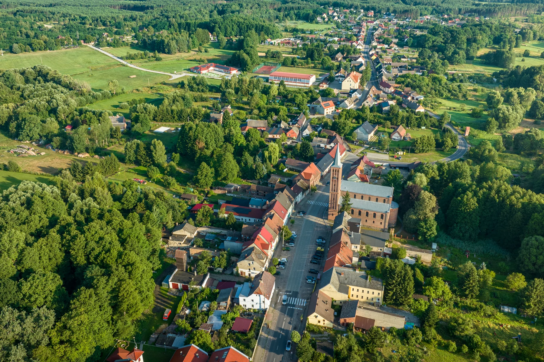Zabudowa ciągnąca się po obu stronach długiej drogi, pośrodku kościół, wokół lasy i pola, widok z lotu ptaka