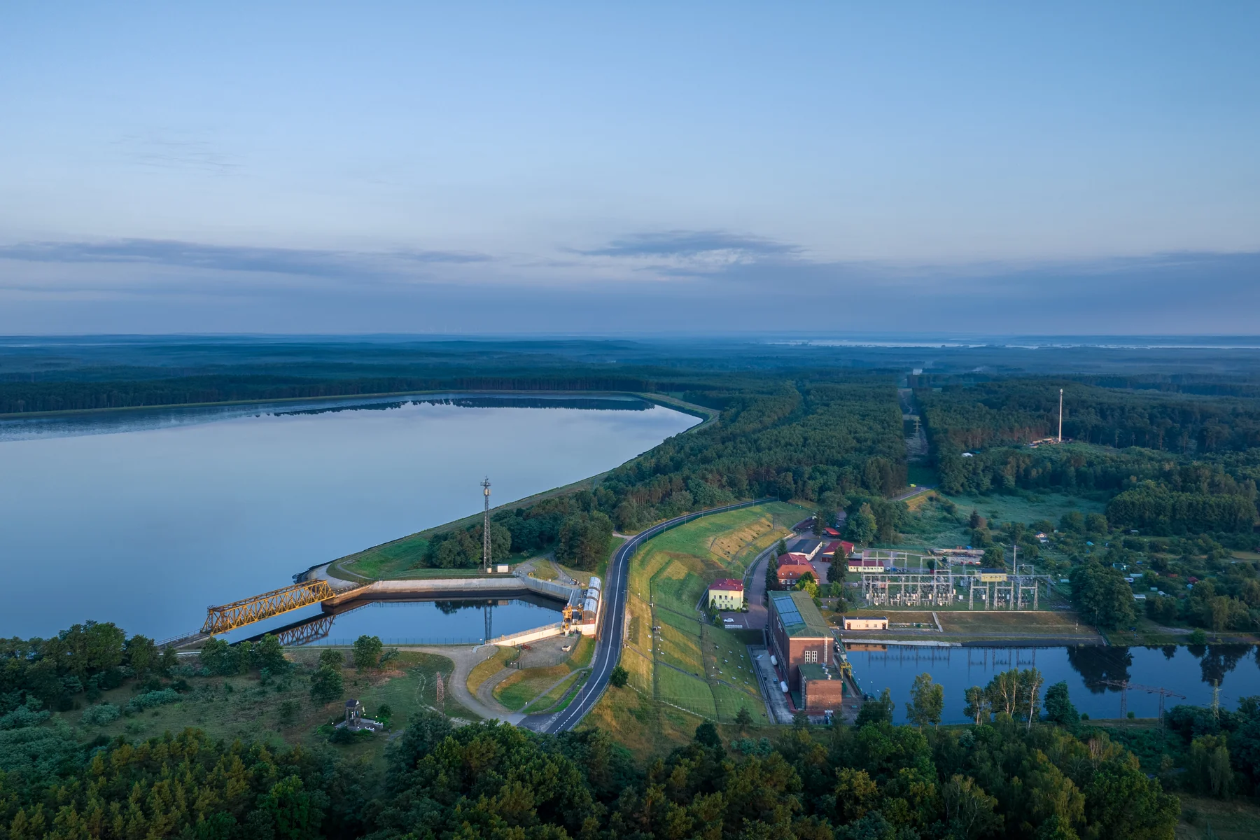 Zbiornik elektrowni wodnej w Dychowie z obwałowaniem i drogą po koronie, u jego stóp budowle hydrotechniczne i stacja elektroenergetyczna, widok z lotu ptaka